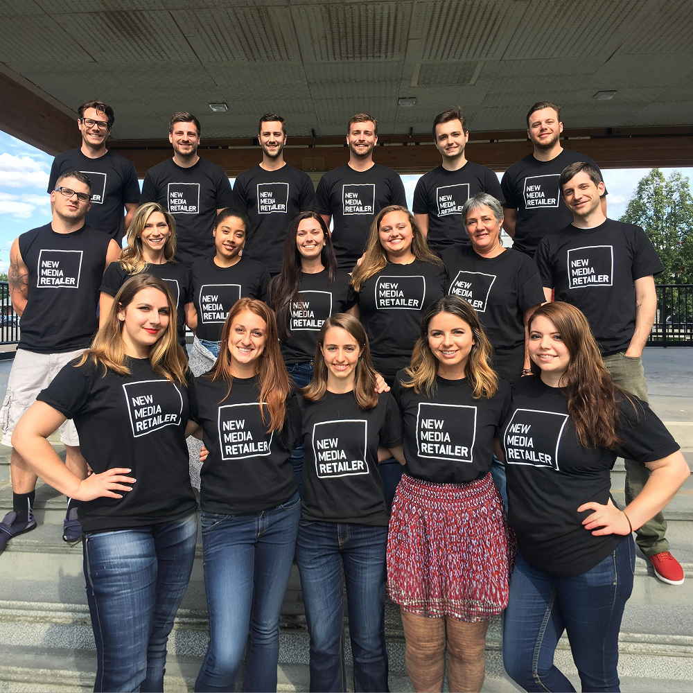 Group of people wearing black t-shirts with 'New Media Retailer' text, standing outdoors.