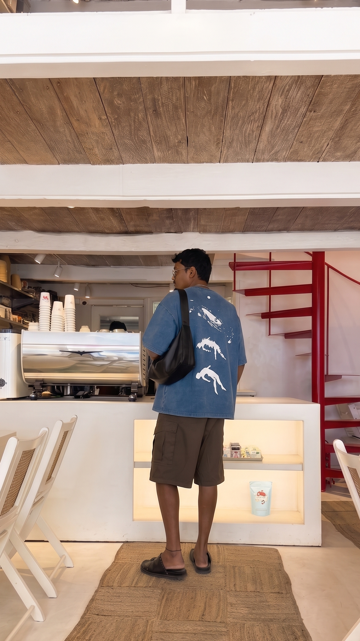 A man wearing a Scrollfit blue graphic t-shirt, brown shorts, and a black shoulder bag stands in a cafe, facing a counter with an espresso machine and shelves.
