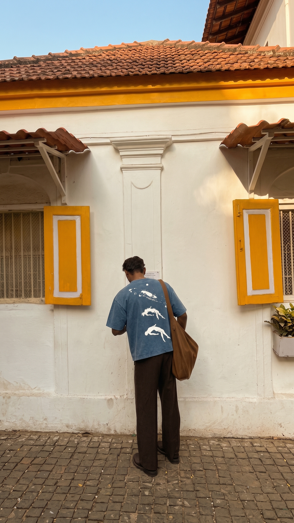 A man stands with his back to the camera wearing a Scrollfit blue denim t-shirt with a white graphic print on the back, paired with dark brown trousers and a brown shoulder bag, posing in front of a white wall with yellow shutters.