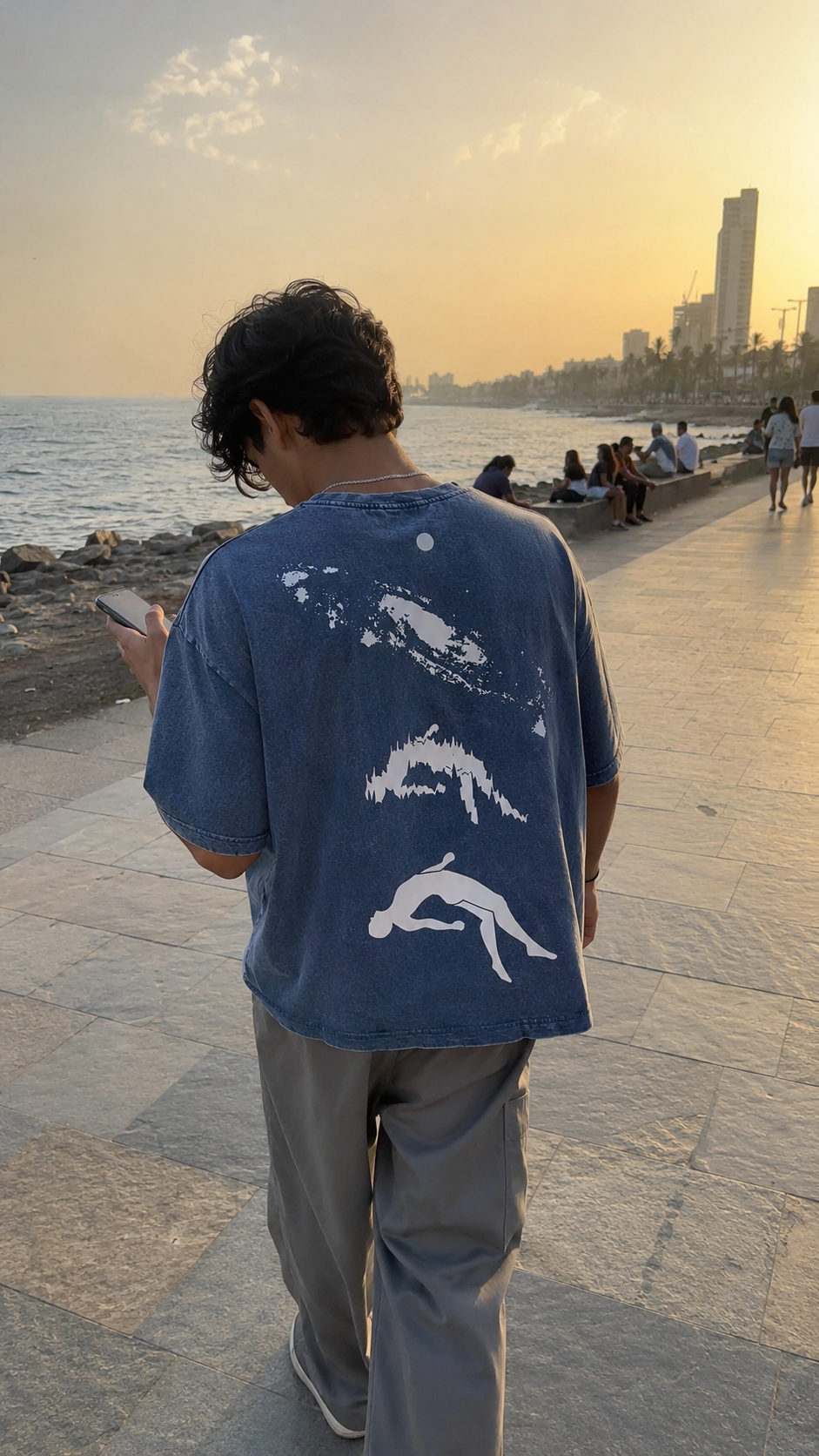 A man viewed from behind walking on a seaside promenade at sunset, wearing a Scrollfit faded blue graphic T-shirt with white shark designs on the back and gray pants.