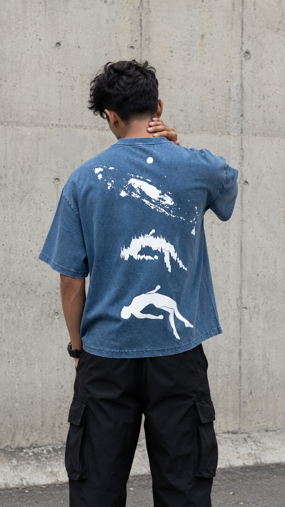 A man viewed from behind wearing a Scrollfit washed blue t-shirt with a large white graphic print of three diving figures on the back, paired with black cargo pants, standing against a concrete wall.