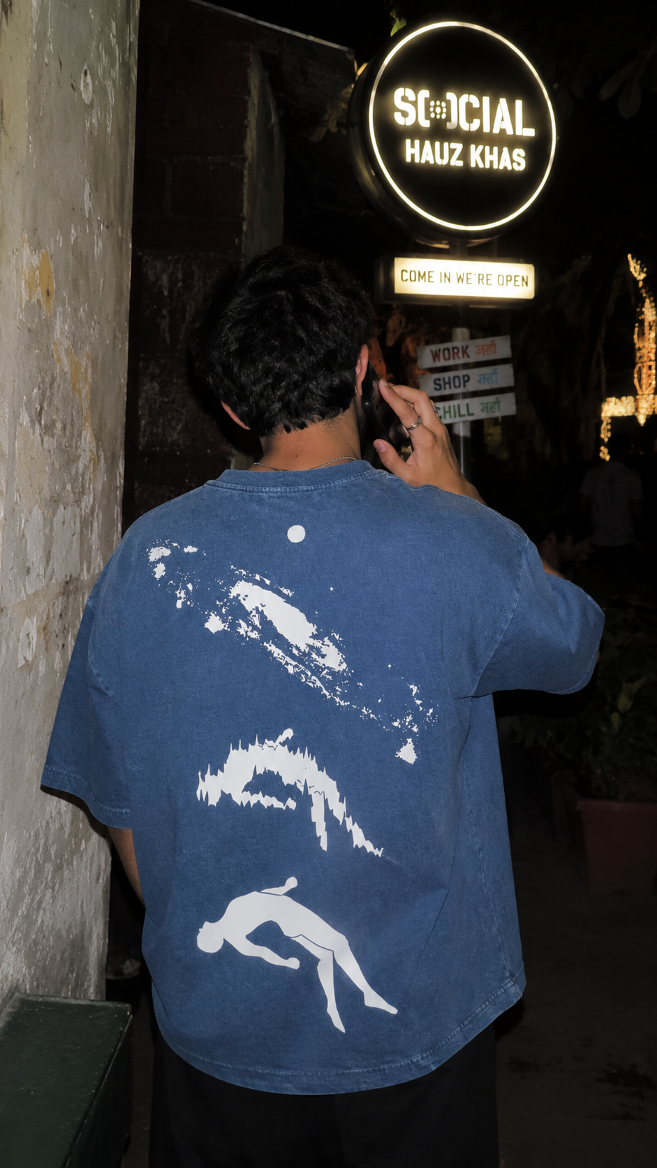 A man seen from behind wearing a blue Scrollfit oversized t-shirt with a white graphic print on the back, standing in a dimly lit outdoor setting near a sign that reads 'St:cial HAUZ KHAS'.