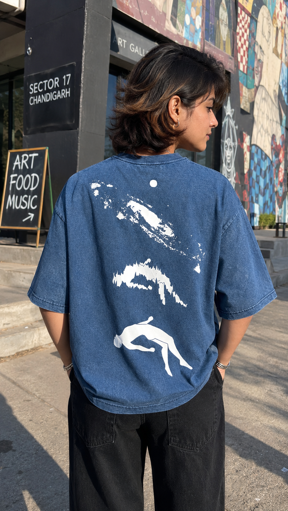 A person viewed from behind wearing an Scrollfit oversized blue graphic T-shirt with white abstract and human figure prints on the back, standing outdoors in front of a graffiti-covered wall.