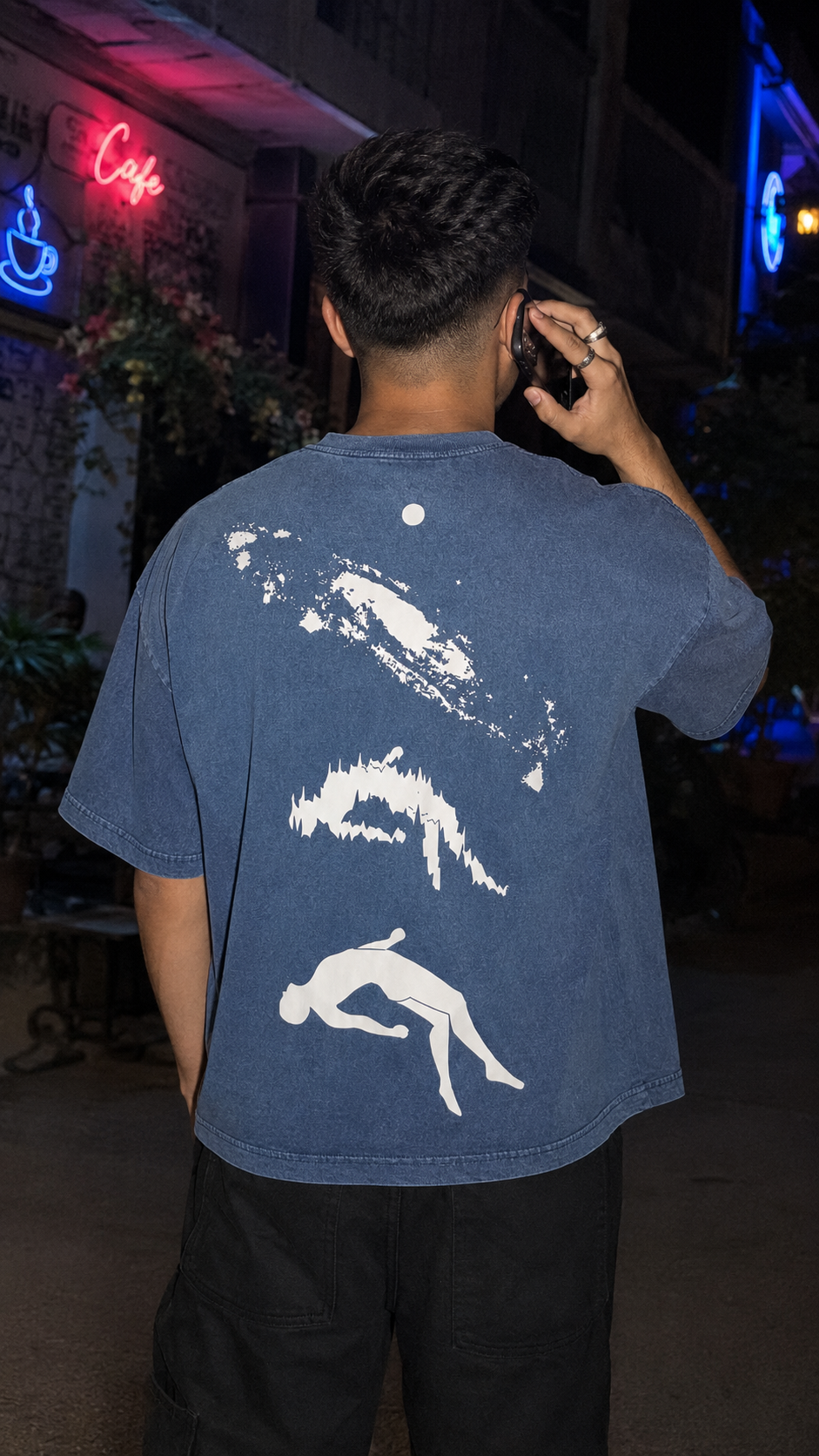 A man viewed from behind wearing a Scrollfit faded blue t-shirt with a white graphic print of three stylized figures on the back, set against a dark, nighttime street cafe background.