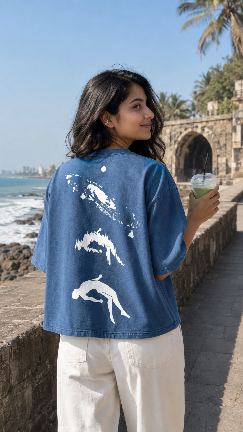 A woman stands outdoors by the sea wearing a Scrollfit blue graphic T-shirt with white abstract designs on the back and white pants, holding a drink in one hand.