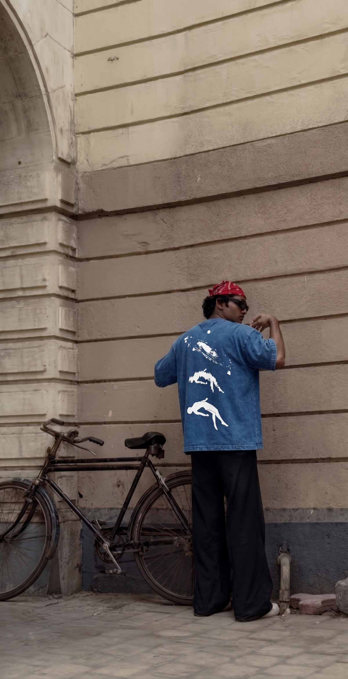 A person stands with their back to the camera wearing a Scrollfit blue denim jacket with white shark graphics, black pants, and a red bandana, standing next to a vintage bicycle against a beige stone wall.