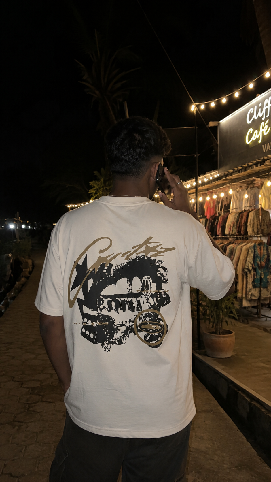 A man viewed from behind wearing a Scrollfit white t-shirt with a large black graphic print on the back, standing outdoors at night next to a shop.