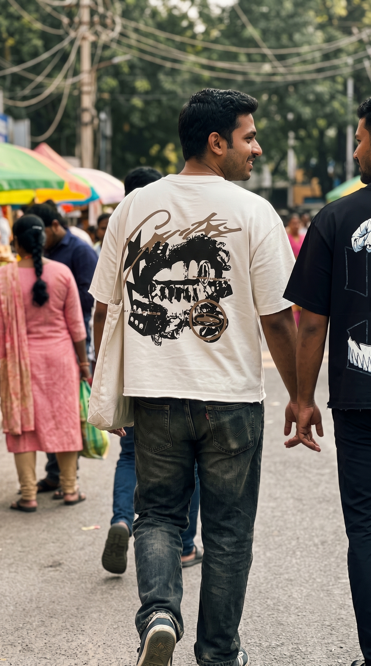 A man walking away in a crowded outdoor street scene, wearing a Scrollfit cream-colored t-shirt with a large black graphic print on the back and dark jeans.