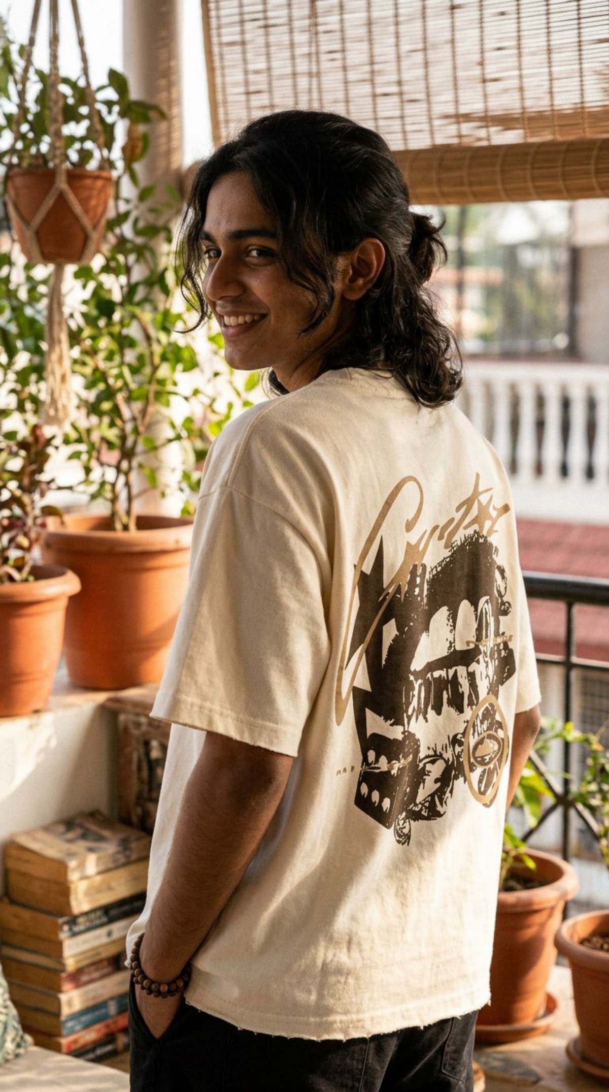A man wearing a Scrollfit cream-colored graphic t-shirt with a large brown motorcycle print on the back, standing in a sunlit room with potted plants and books.