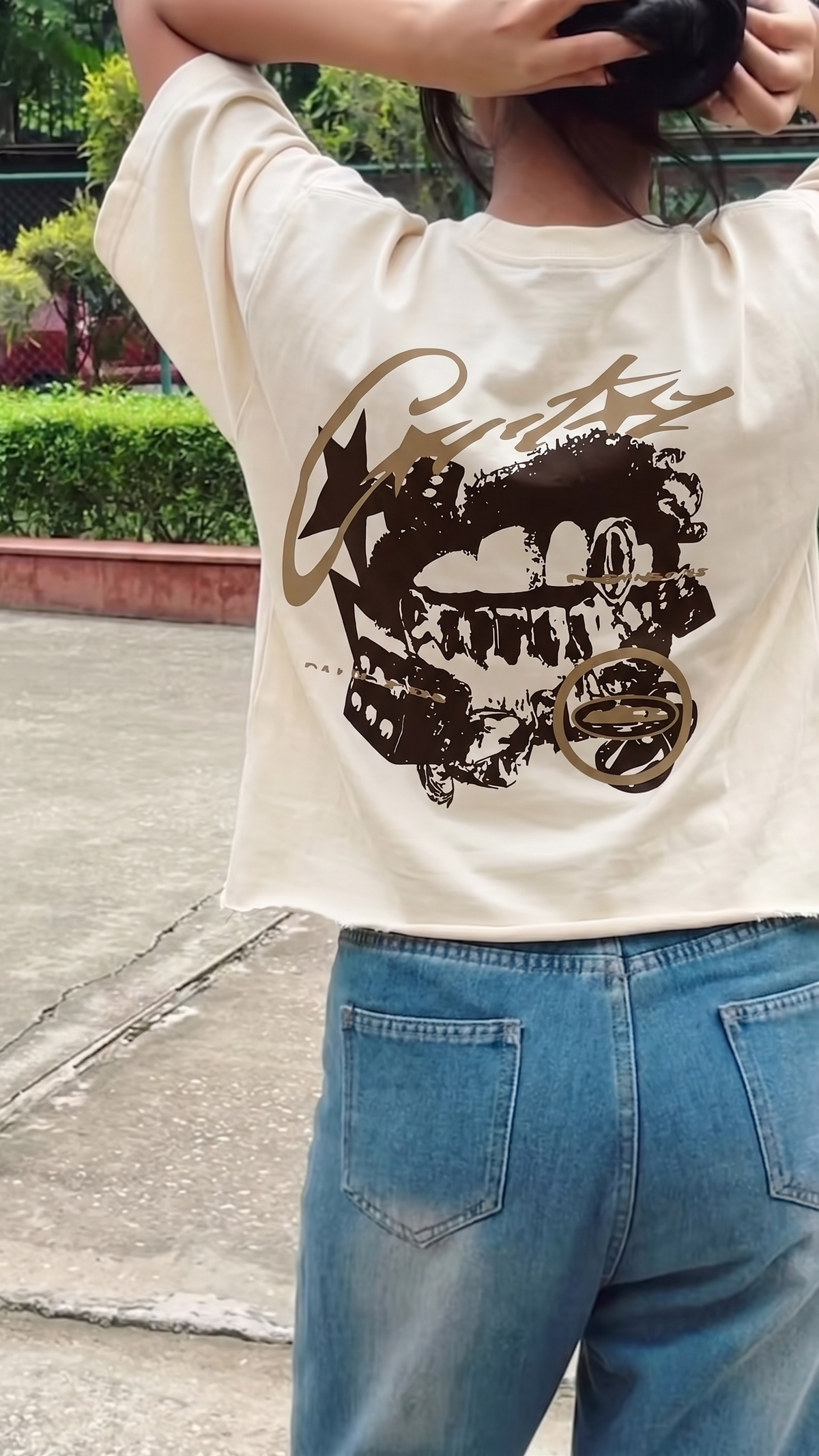 A person wearing a Scrollfit cream-colored graphic t-shirt with a large black and brown skull design and blue jeans, photographed outdoors.