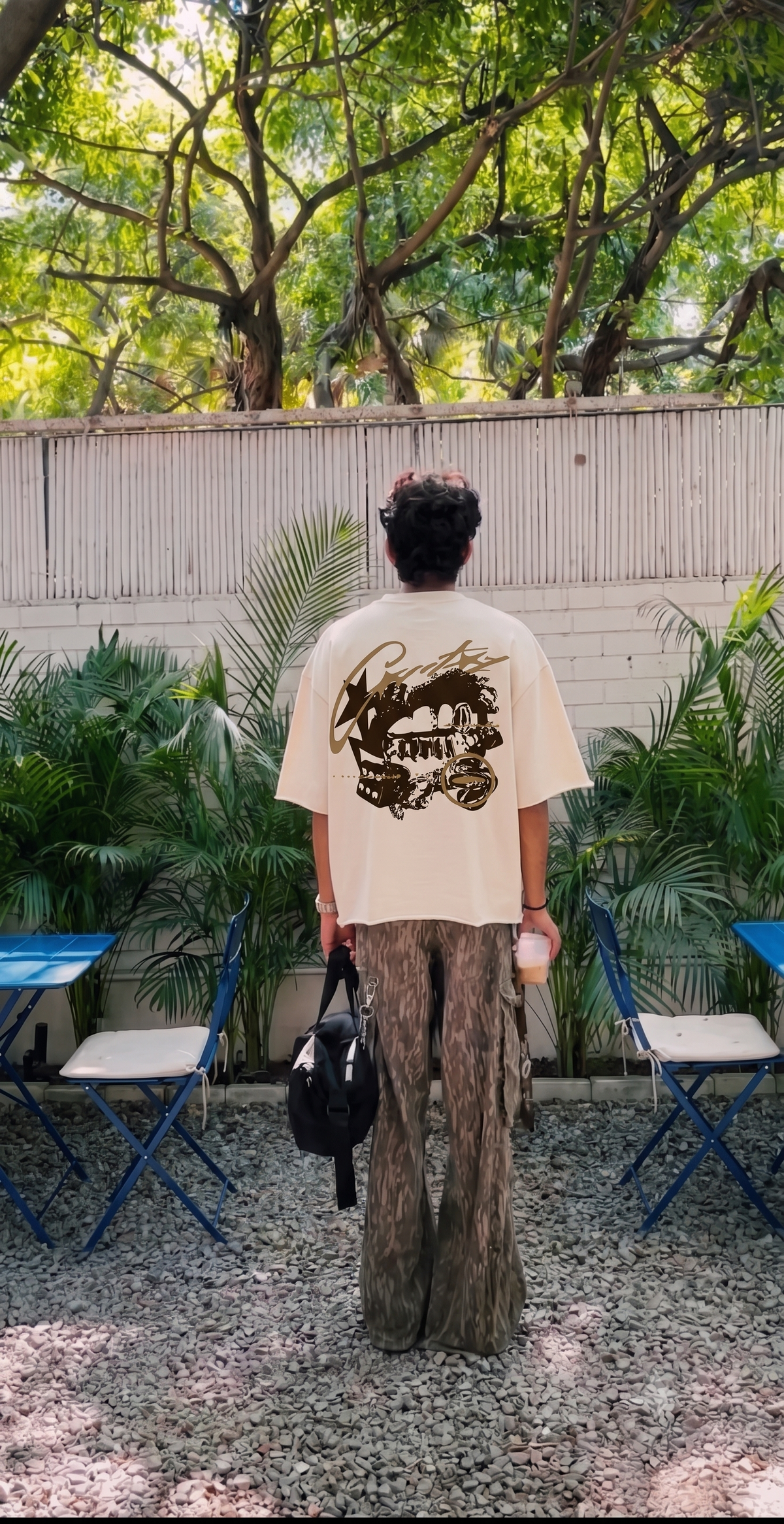A person standing outdoors in a backyard patio setting, viewed from behind, wearing a Scrollfit cream-colored oversized t-shirt with a large black graphic print on the back and wide-leg brown pants.
