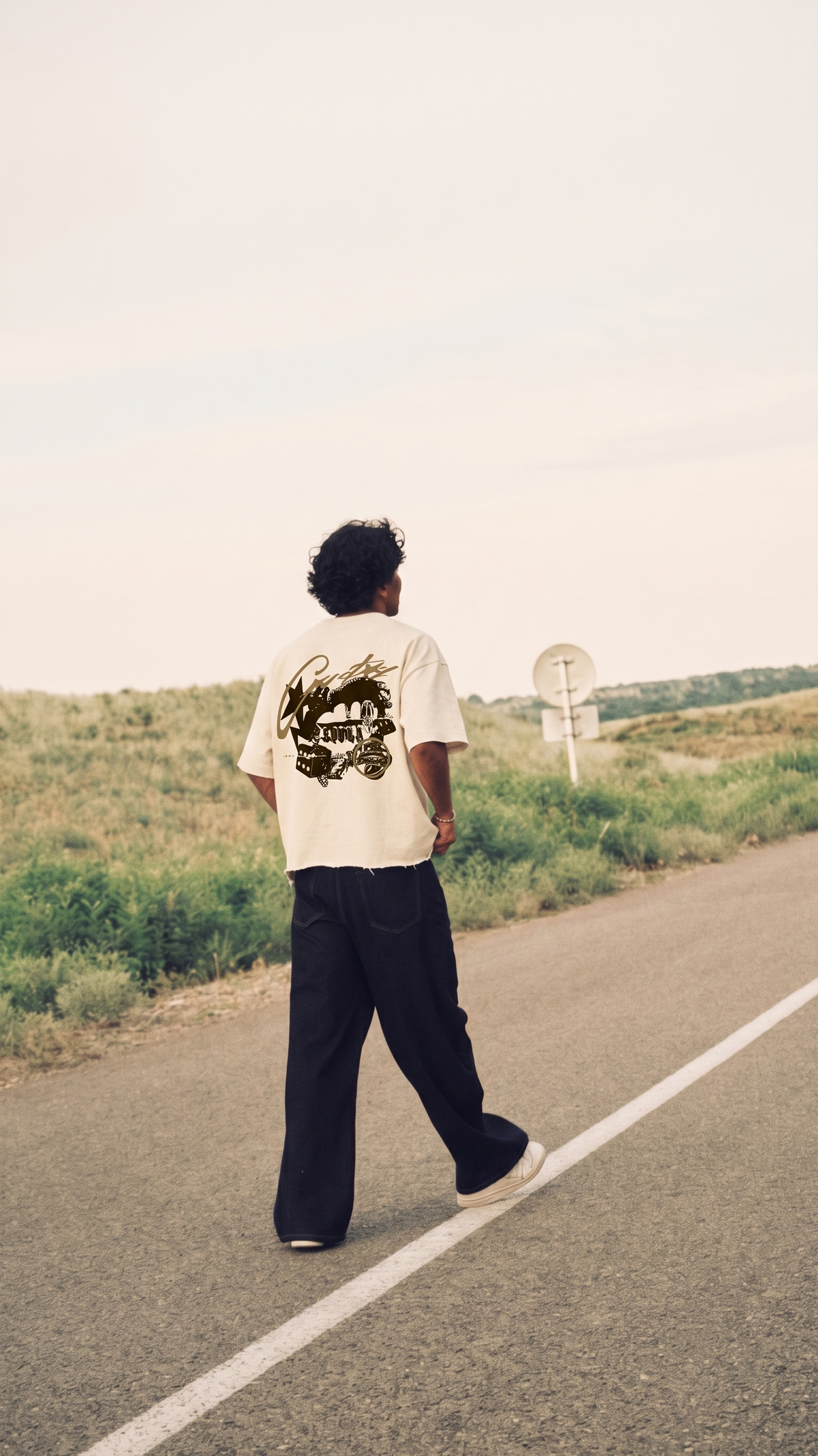 A man stands on a paved road in a rural landscape, viewed from behind, wearing a Scrollfit cream-colored t-shirt with a large black graphic print on the back and loose black pants.