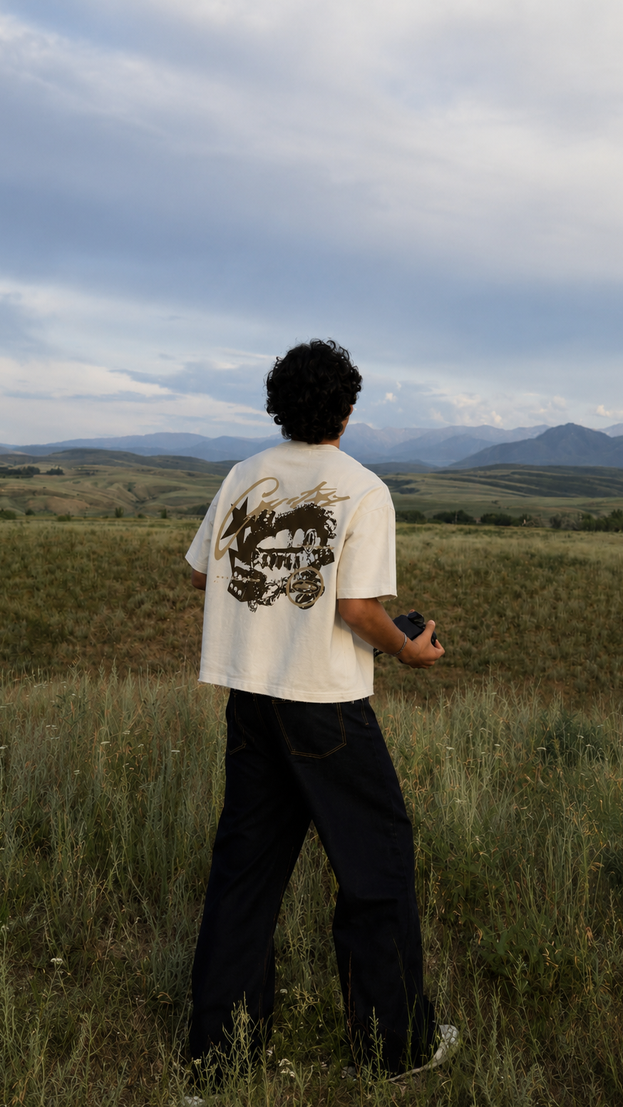 A person standing in a grassy field with mountains and a cloudy sky in the background, viewed from behind. The person is wearing a Scrollfit cream-colored graphic T-shirt with a dark print on the back and loose black pants.