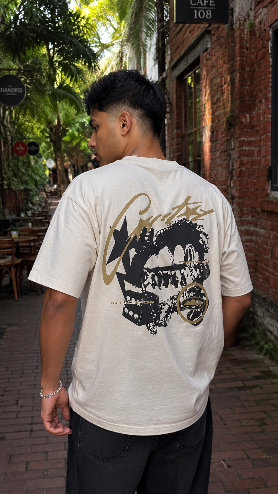 A man viewed from behind wearing a Scrollfit cream-colored t-shirt with a large black and gold graphic print on the back, standing on a brick sidewalk in an outdoor alleyway.