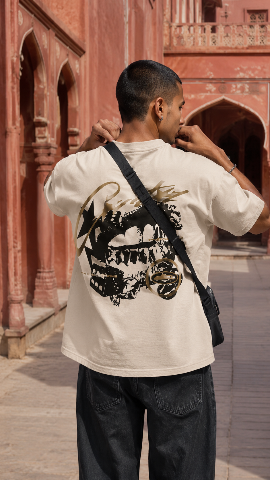 A man viewed from behind wearing a Scrollfit cream-colored t-shirt with a large black and gold graphic print on the back, paired with dark jeans and a black crossbody bag, standing in front of a red sandstone architectural background.