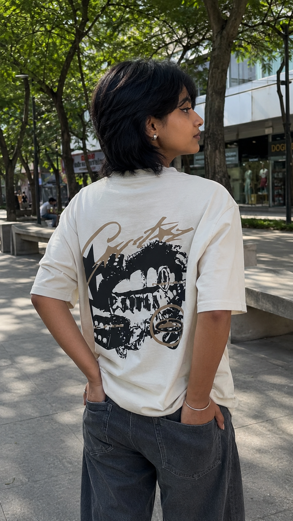 A person standing outdoors on a tree-lined street, wearing a Scrollfit cream-colored graphic t-shirt with a large black and brown skull design on the back, paired with dark gray wide-leg jeans.