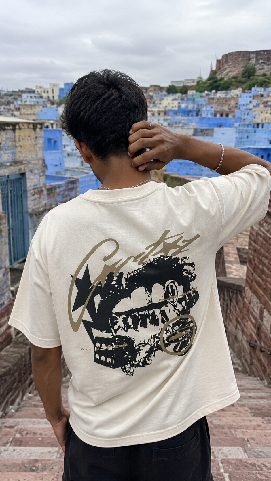 A man viewed from behind wearing a Scrollfit cream-colored t-shirt with a large black graphic print on the back, standing on stone steps with a scenic blue-roofed city in the background.