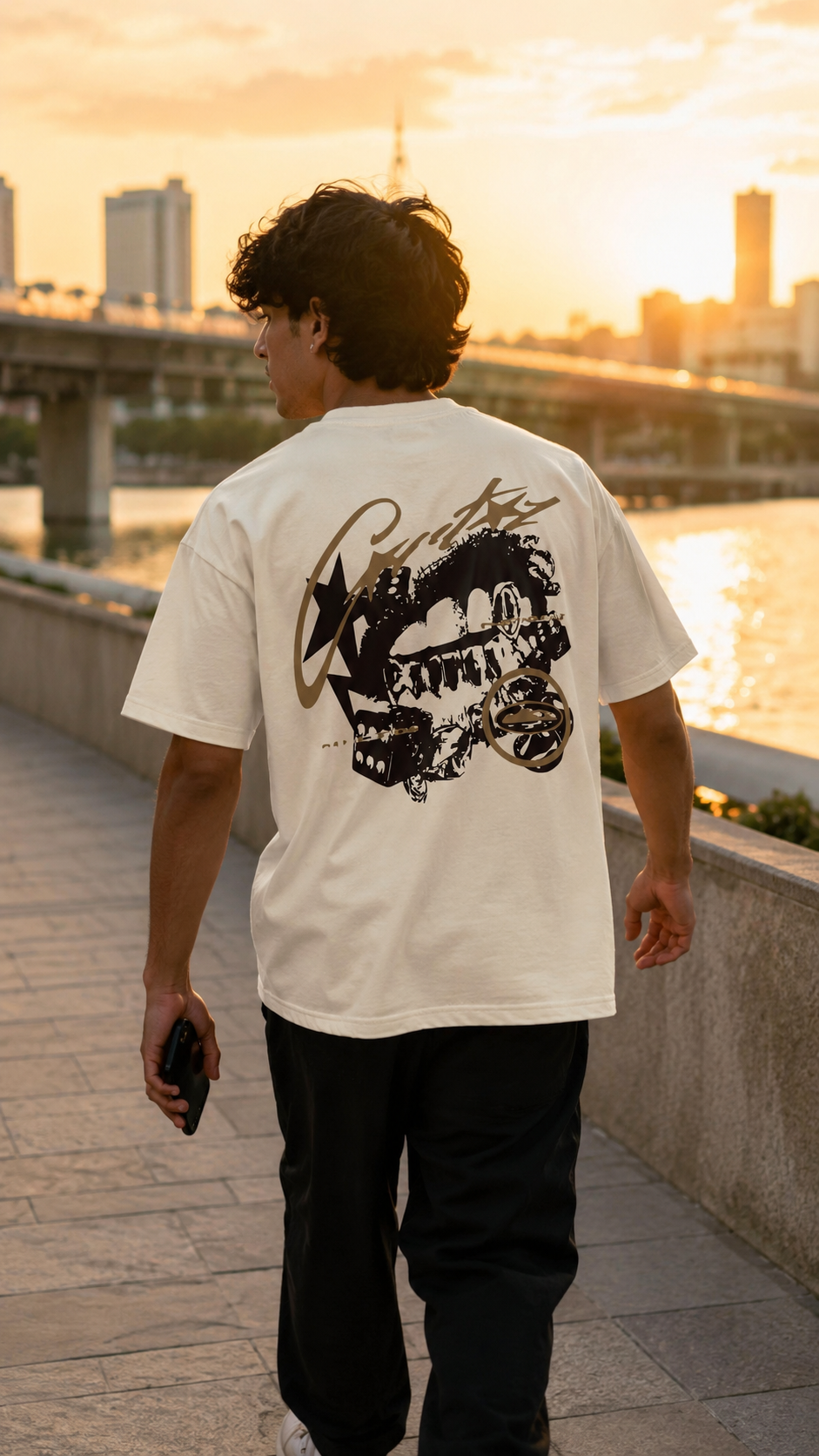 A man viewed from behind walking on a riverside path at sunset, wearing a Scrollfit white short-sleeve T-shirt with a large black graphic print on the back and black pants.