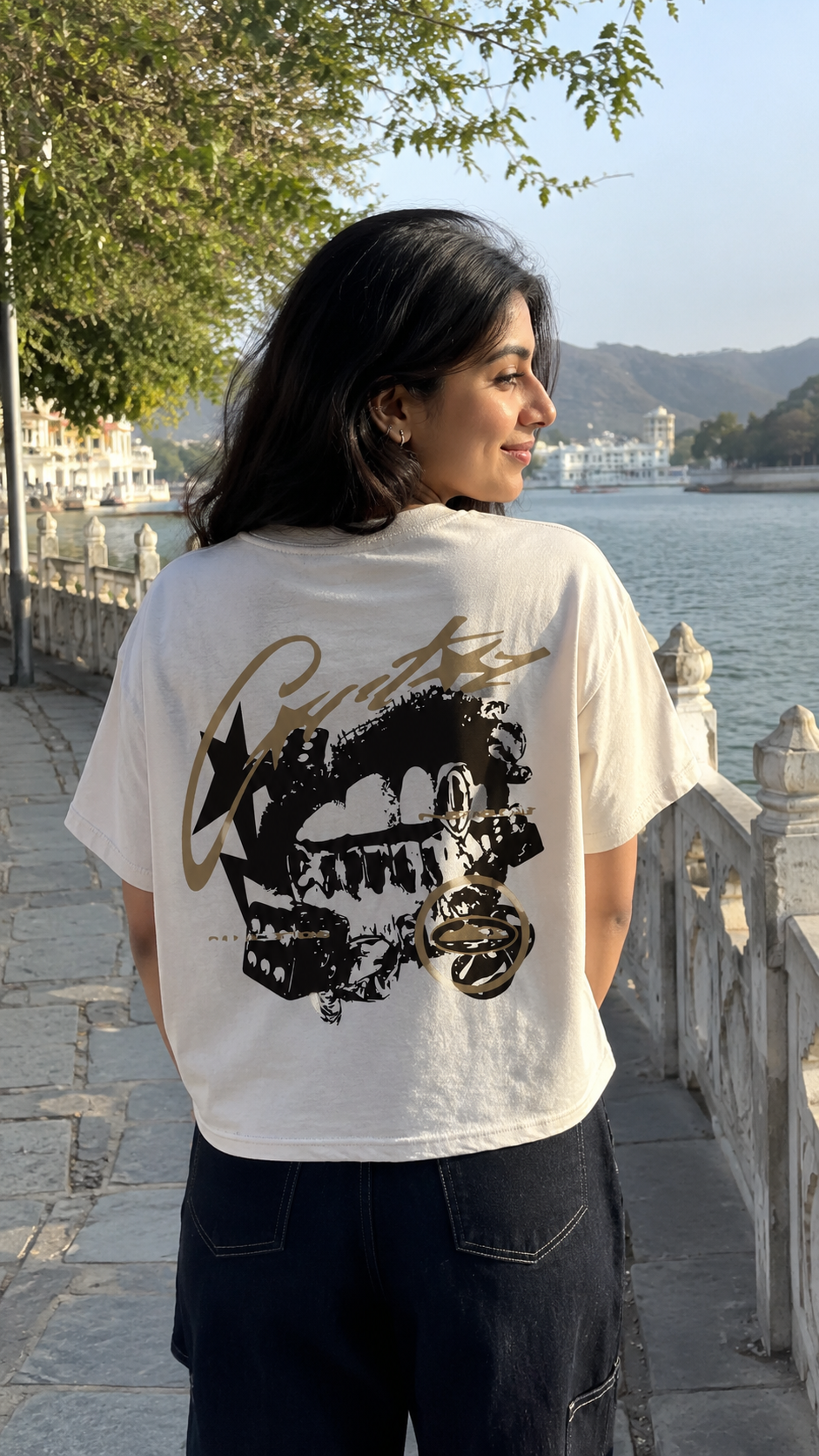 A woman standing outdoors by a lake wearing a Scrollfit off-white graphic T-shirt with a large black and gold print on the back, paired with dark jeans.