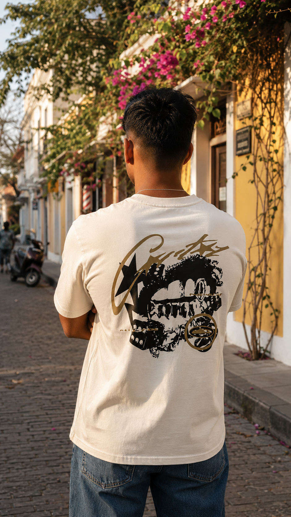 A man viewed from behind wearing a Scrollfit cream-colored t-shirt with a large black and gold graphic print on the back, standing on a cobblestone street in a sunny, European-style town.