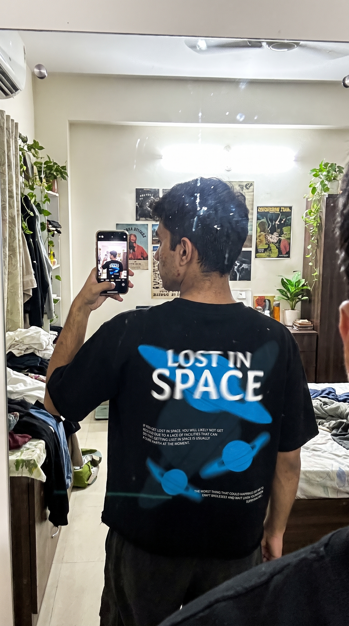 A man taking a mirror selfie in a bedroom, wearing a Scrollfit black t-shirt with a blue and white graphic that reads 'LOST IN SPACE'.