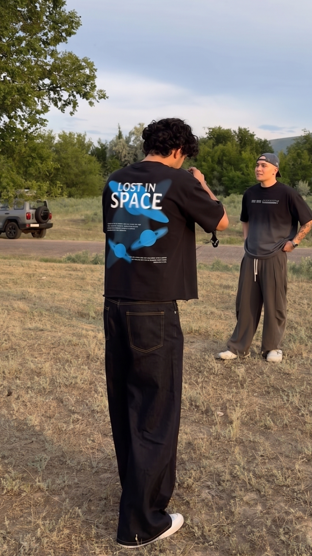 A man wearing a Scrollfit black graphic t-shirt with 'LOST IN SPACE' and blue space-themed graphics on the back, standing in an outdoor grassy field. Another man is visible in the background wearing a Scrollfit dark gray t-shirt and sweatpants.