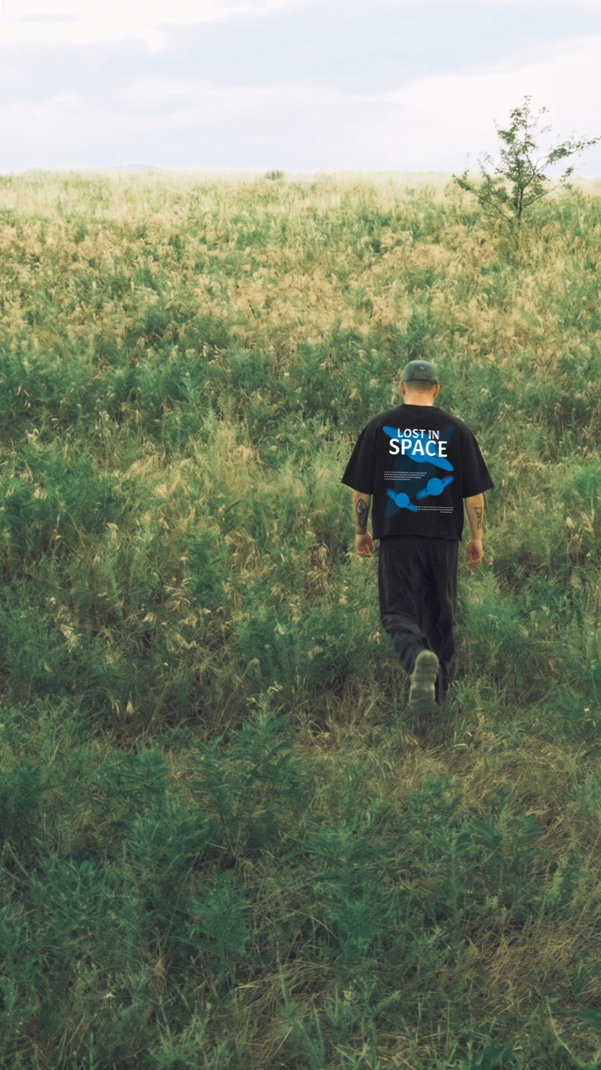 A man walking through a grassy field, viewed from behind, wearing a Scrollfit black t-shirt with a blue graphic and the words 'LOST IN SPACE' printed on the back.