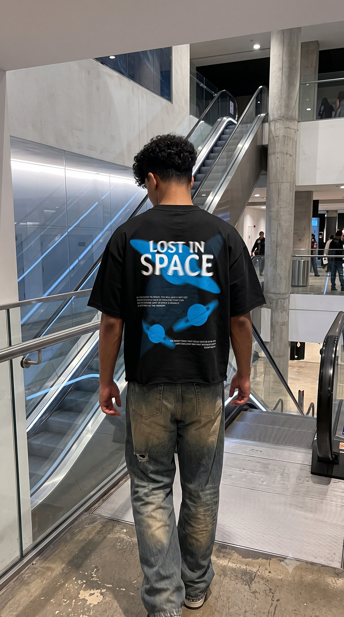 A man viewed from behind wearing a Scrollfit black graphic t-shirt with 'LOST IN SPACE' and blue space-themed graphics, paired with faded gray jeans, standing in a modern indoor escalator area.