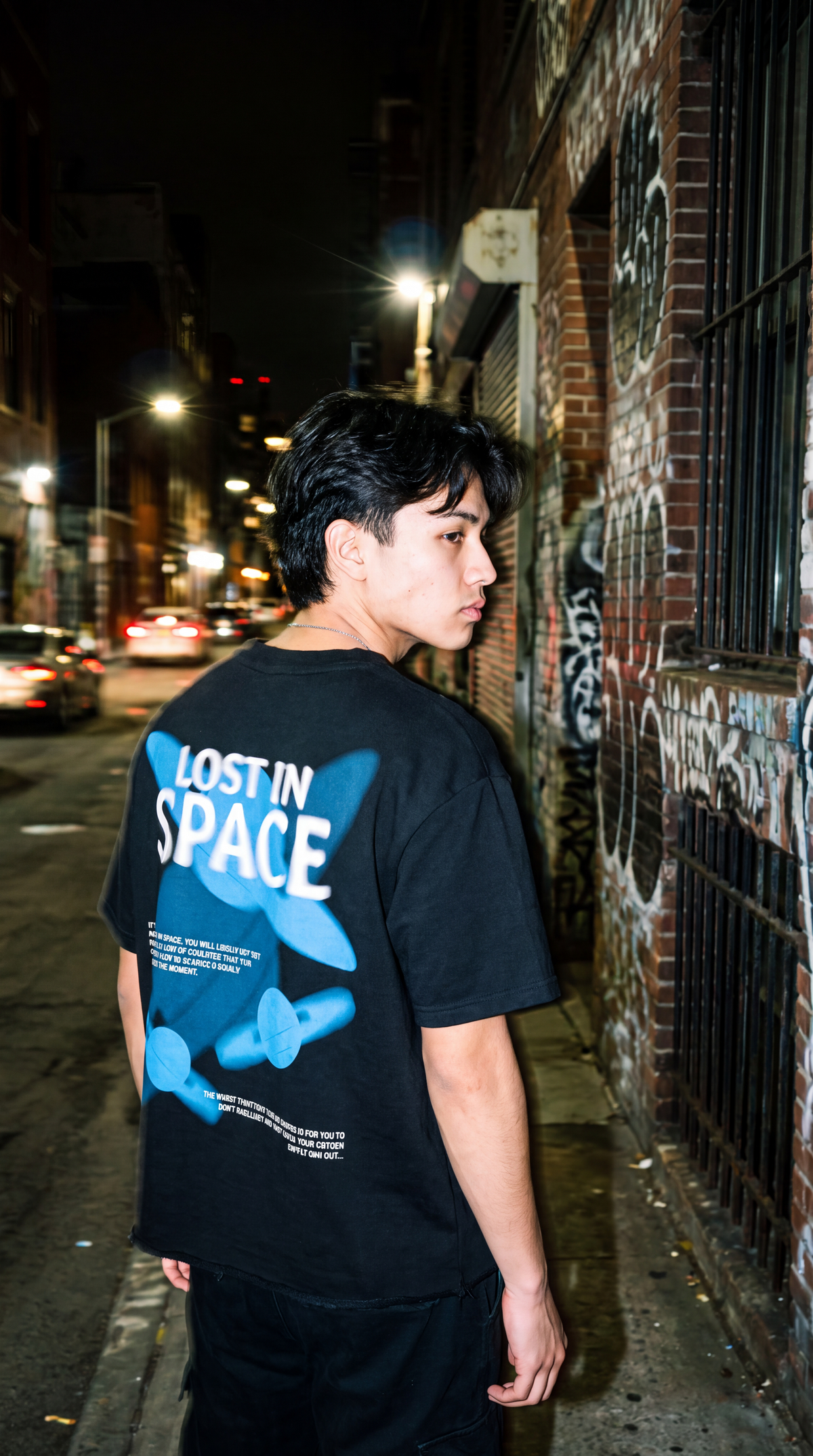 A man standing on a city street at night wearing a Scrollfit black t-shirt with a large blue and white graphic on the back that reads 'LOST IN SPACE'.
