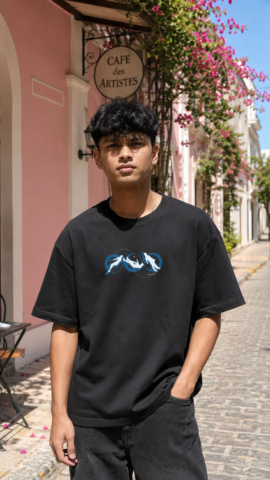 A man wearing a Scrollfit black graphic t-shirt with a blue and white shark design, standing on a cobblestone street in front of a pink building with a cafe sign.