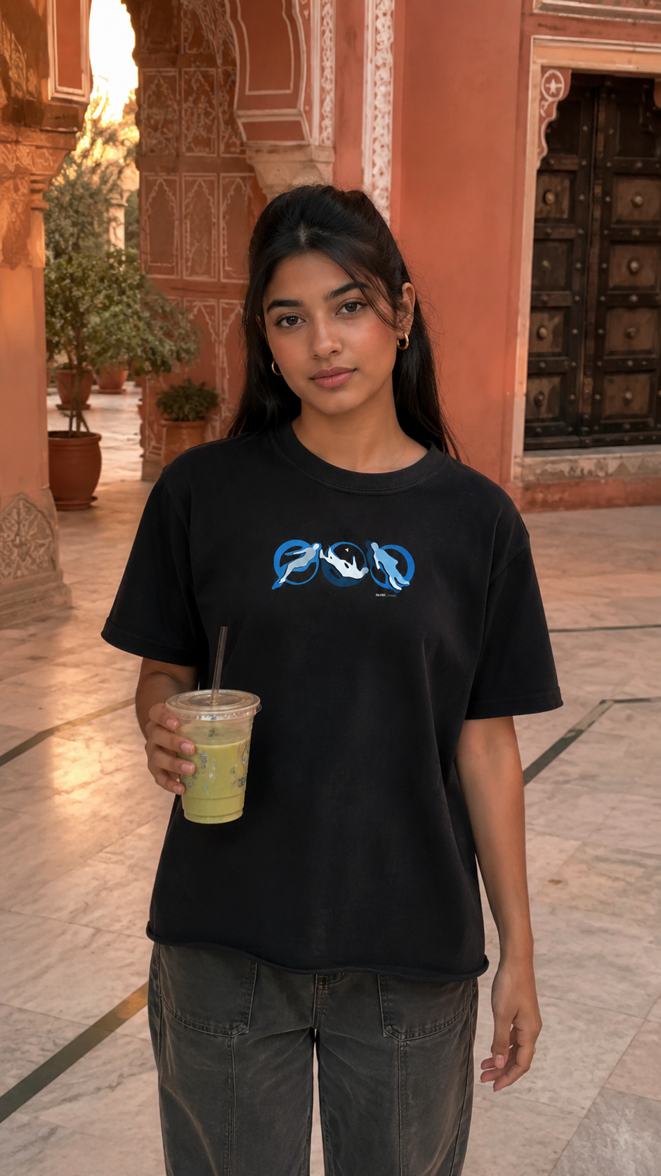 A woman wearing a Scrollfit black t-shirt with a blue and white graphic print, holding a green drink in a courtyard setting with warm-toned walls.