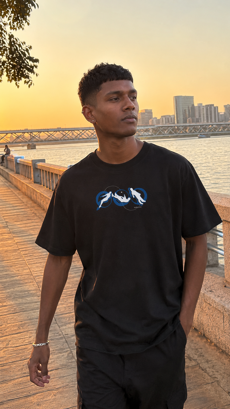 A man wearing a Scrollfit black t-shirt with a blue and white graphic print, standing on a waterfront boardwalk at sunset.