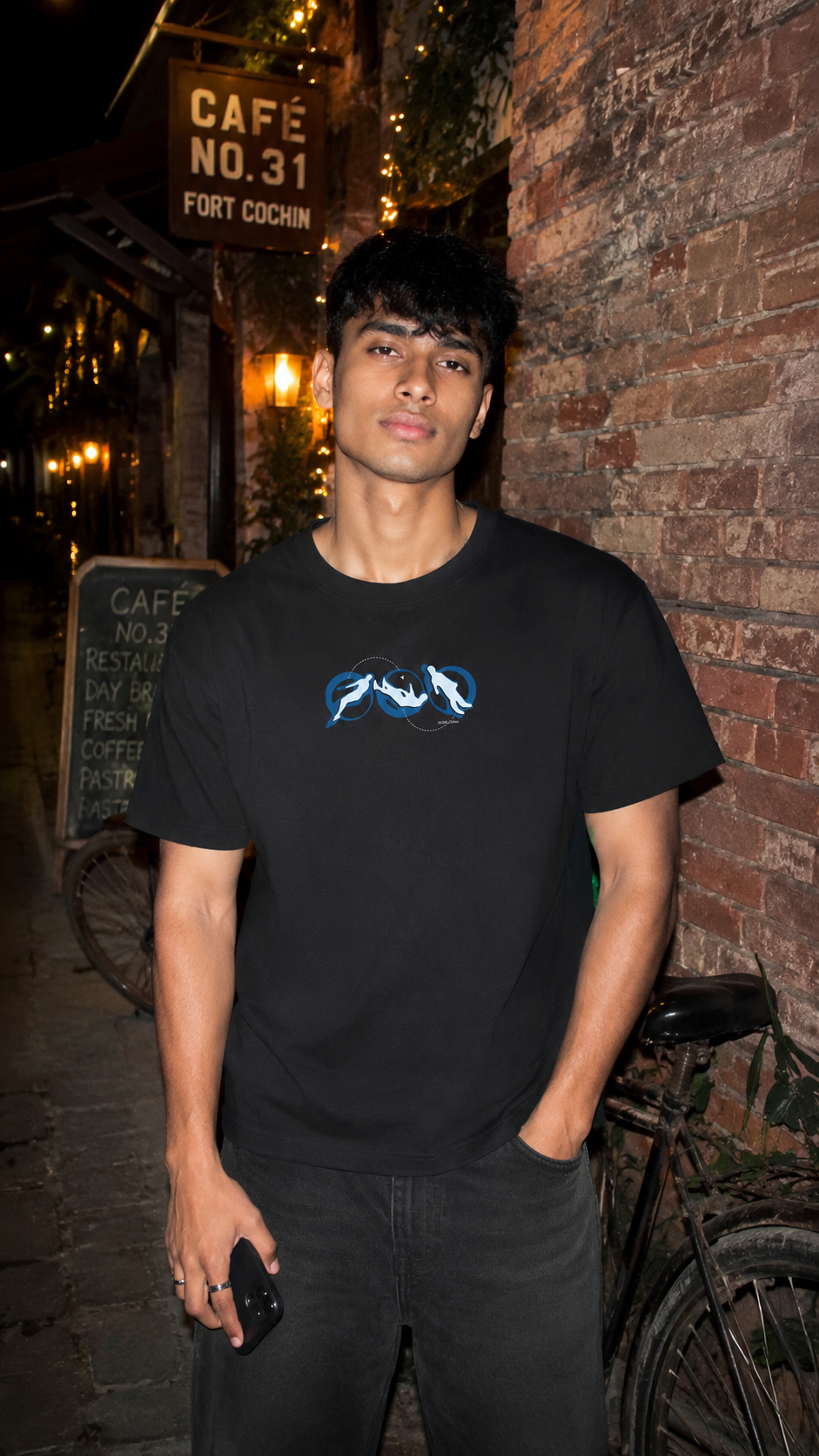 A man wearing a Scrollfit black short-sleeve t-shirt with a blue and white mountain graphic on the chest, standing in front of a brick wall at night.