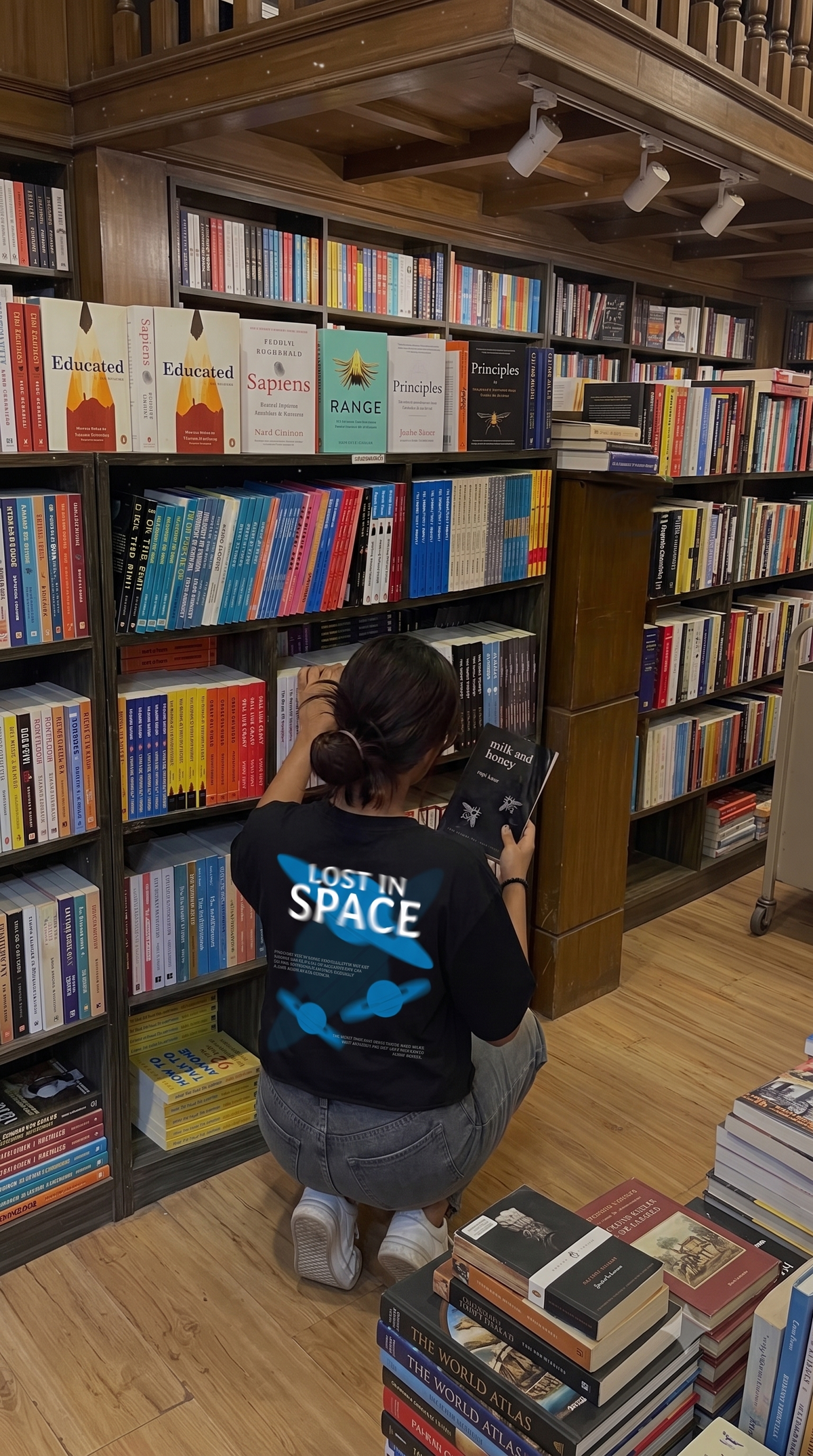 A person crouching in a bookshop, reading a book. They are wearing a Scrollfit black t-shirt with 'LOST IN SPACE' and blue graphics on the back, paired with gray jeans.