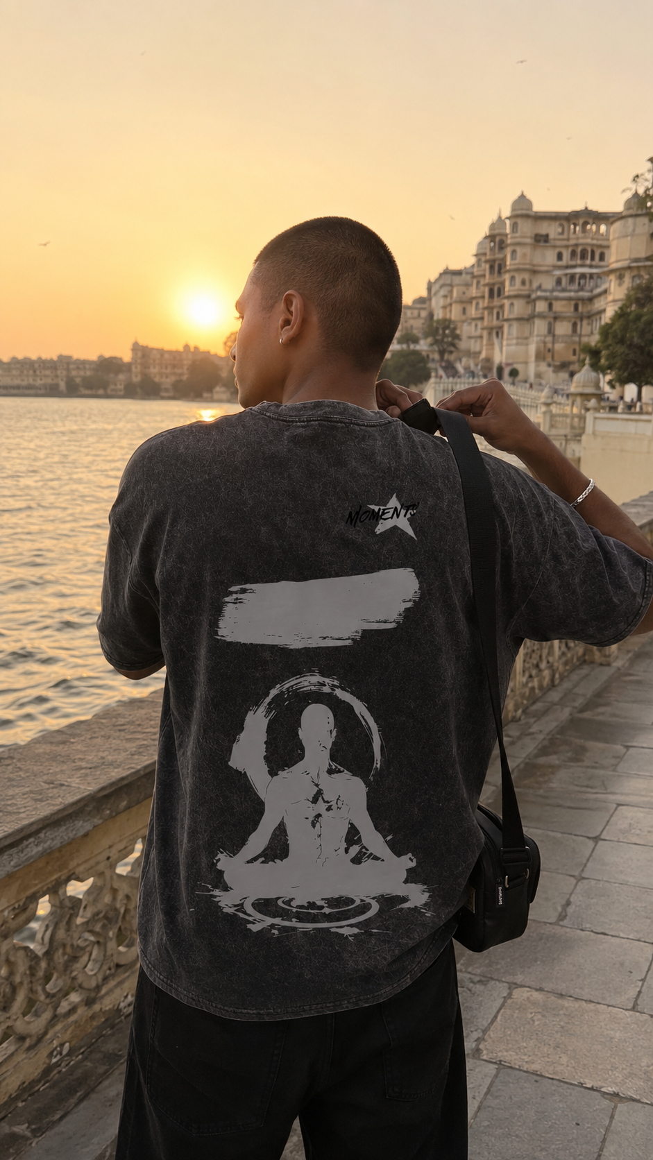 A man seen from behind wearing a Scrollfit dark gray washed t-shirt with a large white graphic of a meditating figure on the back, standing on a riverside walkway at sunset.