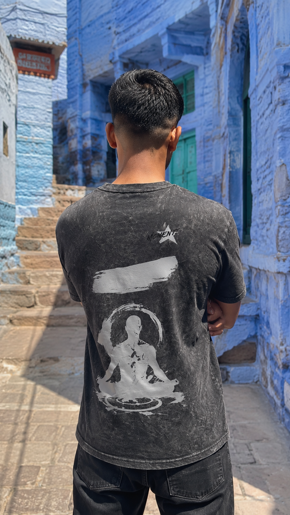 A man viewed from behind wearing a Scrollfit faded black graphic t-shirt with a white meditating figure print on the back, standing in a narrow alleyway with blue stone walls.