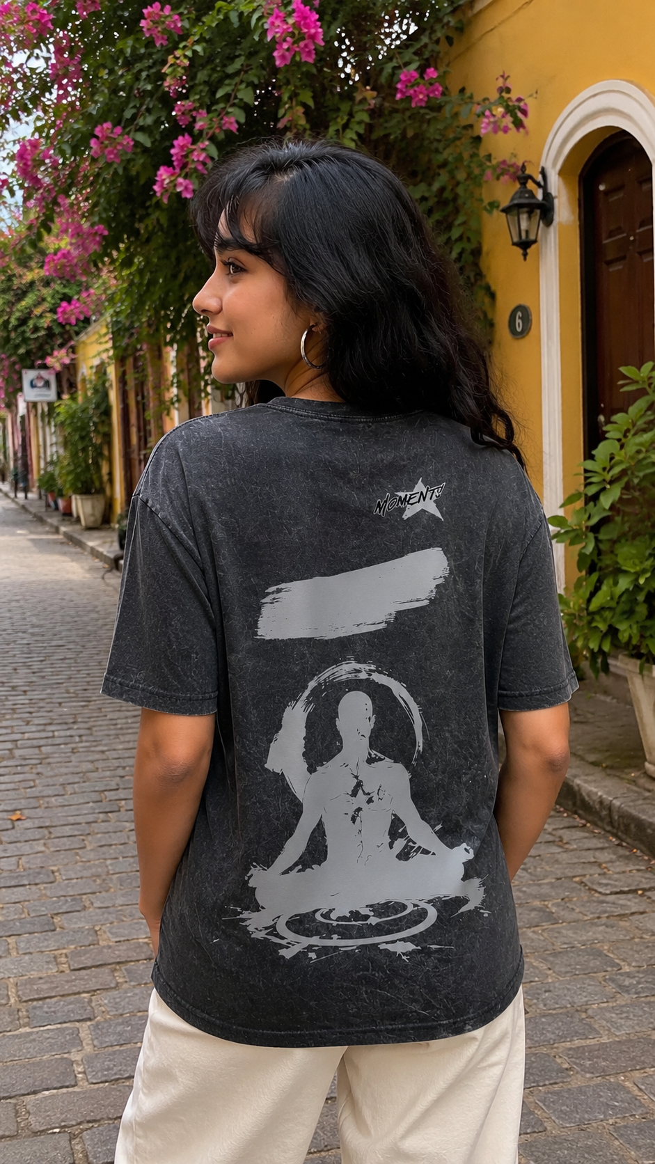 A woman wearing a Scrollfit dark gray mineral-wash graphic t-shirt with a white meditating Buddha design on the back, standing on a cobblestone street in front of a yellow building with pink flowers.