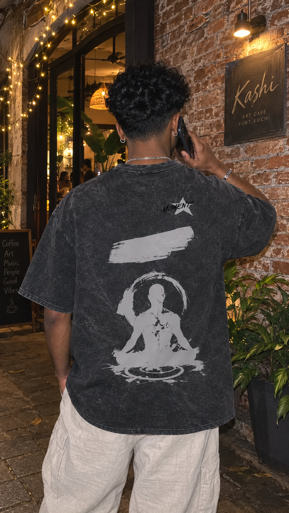 A man viewed from behind wearing a Scrollfit faded black graphic t-shirt with a large white meditating figure design on the back, standing outside a restaurant with brick walls and warm string lights.