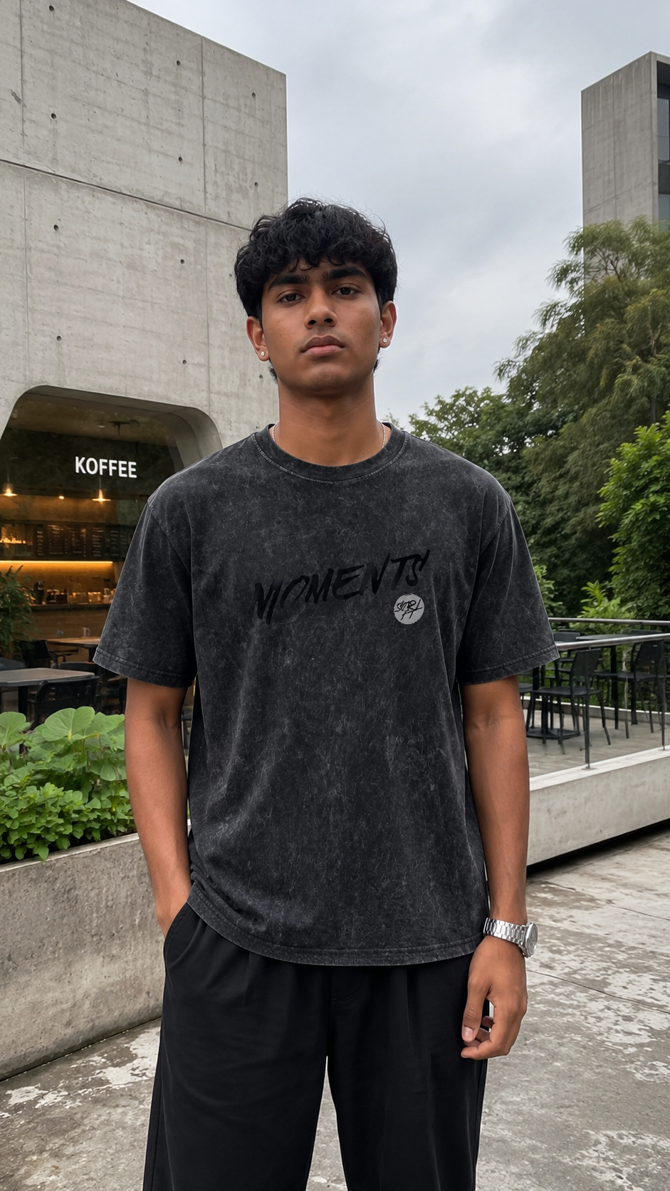 A man wearing a Scrollfit dark gray mineral-wash t-shirt with 'MOMENTS' printed on the front, standing outdoors in front of a concrete building.
