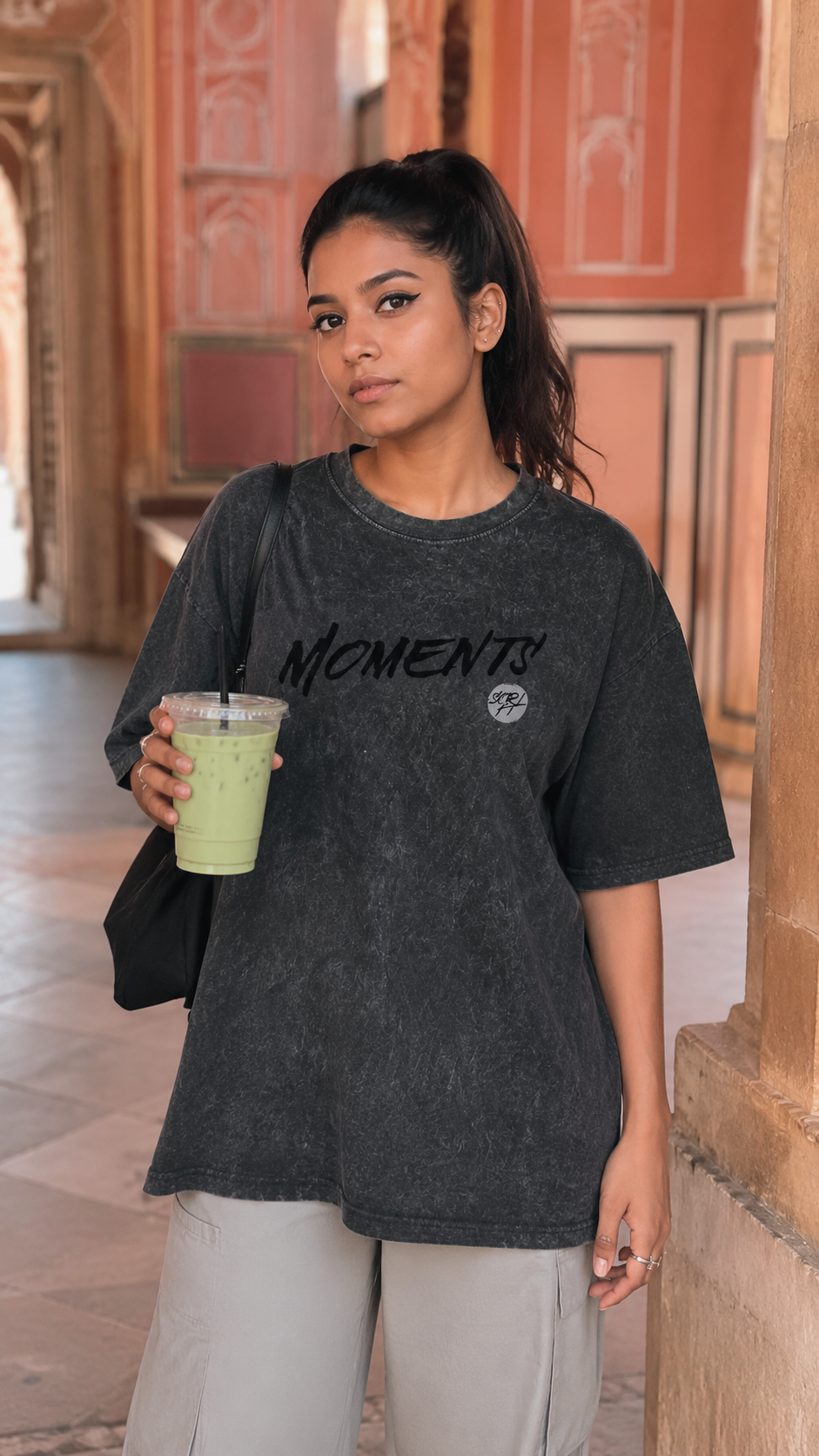 A woman wearing an oversized Scrollfit dark gray washed t-shirt with 'MOMENTS' printed in black across the chest, holding a green drink in a corridor with warm-toned walls.