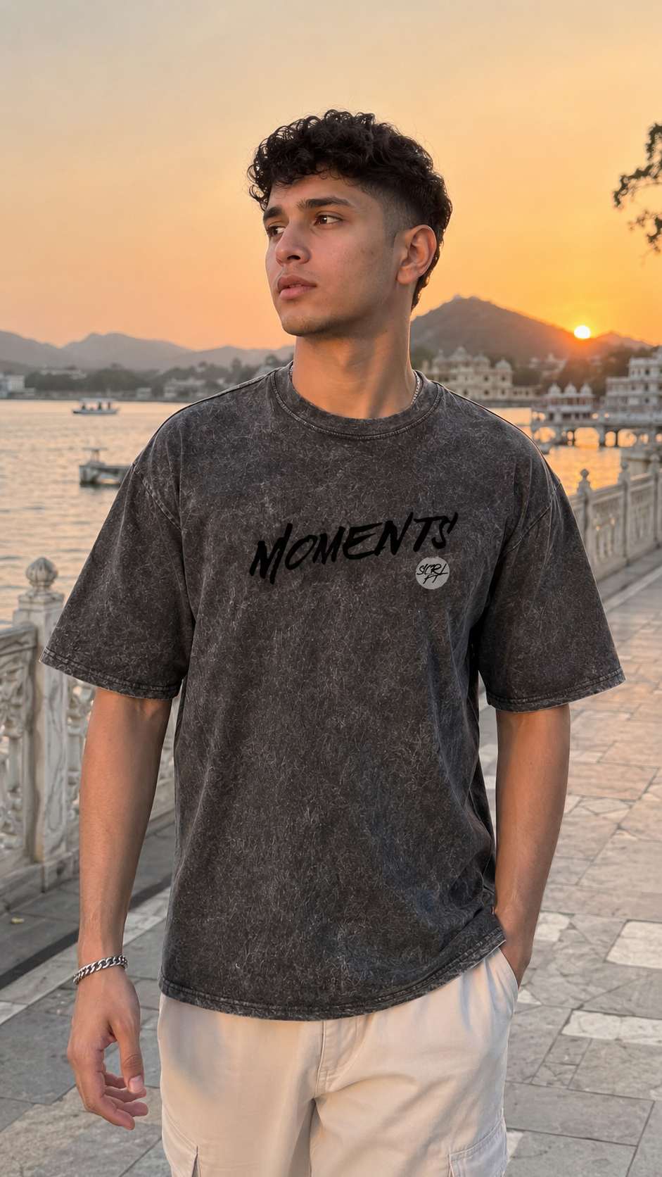 A man wearing a Scrollfit dark gray acid-wash t-shirt with 'MOMENTS' printed in black across the chest, standing outdoors by a lake at sunset.