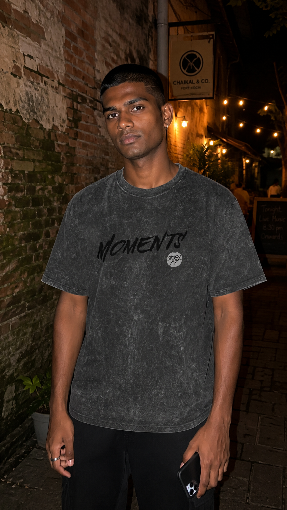 A man wearing a Scrollfit dark gray mineral-wash t-shirt with 'MOMENTS' printed on the chest, standing in a dimly lit outdoor alleyway at night.