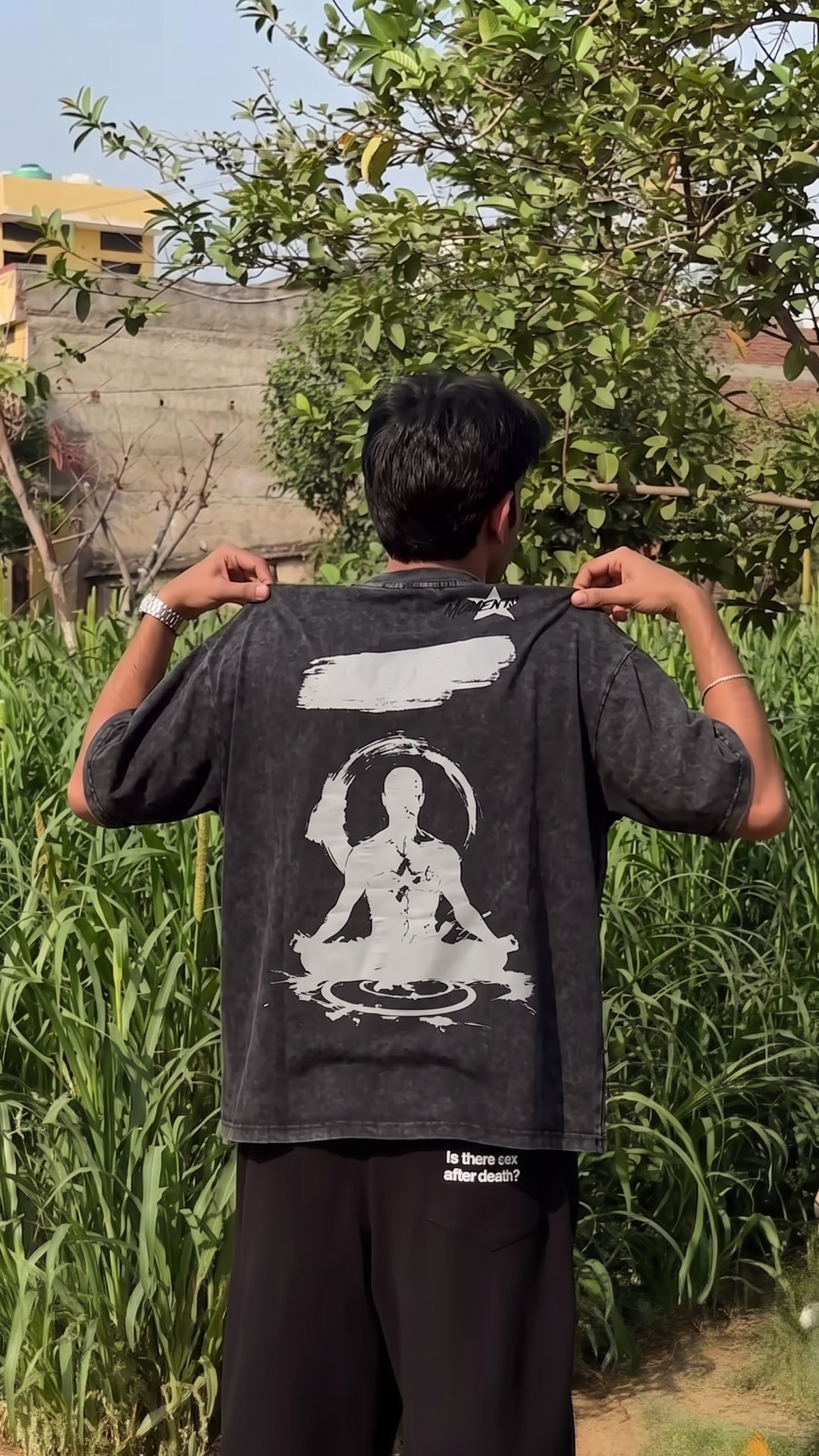 A man viewed from behind wearing a Scrollfit faded black graphic t-shirt with a large white meditating figure print on the back, standing outdoors in a grassy field.