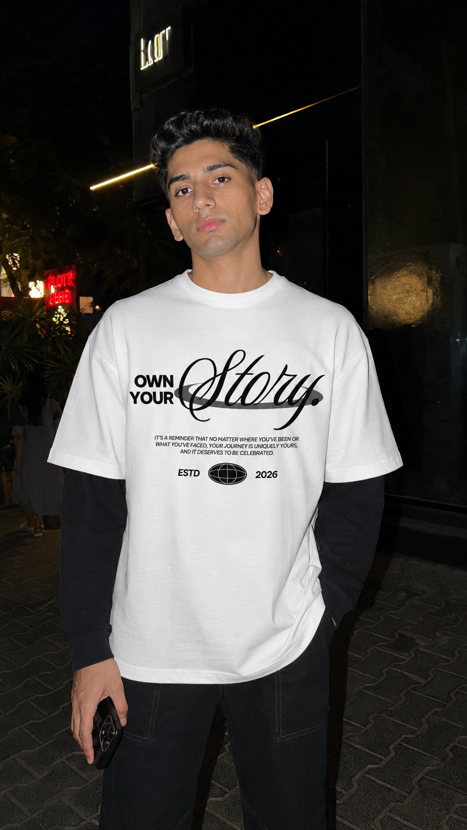A man wearing a Scrollfit white graphic t-shirt with 'OWN YOUR Story' printed on the front, layered over a black long-sleeve shirt and black pants, standing outdoors at night.