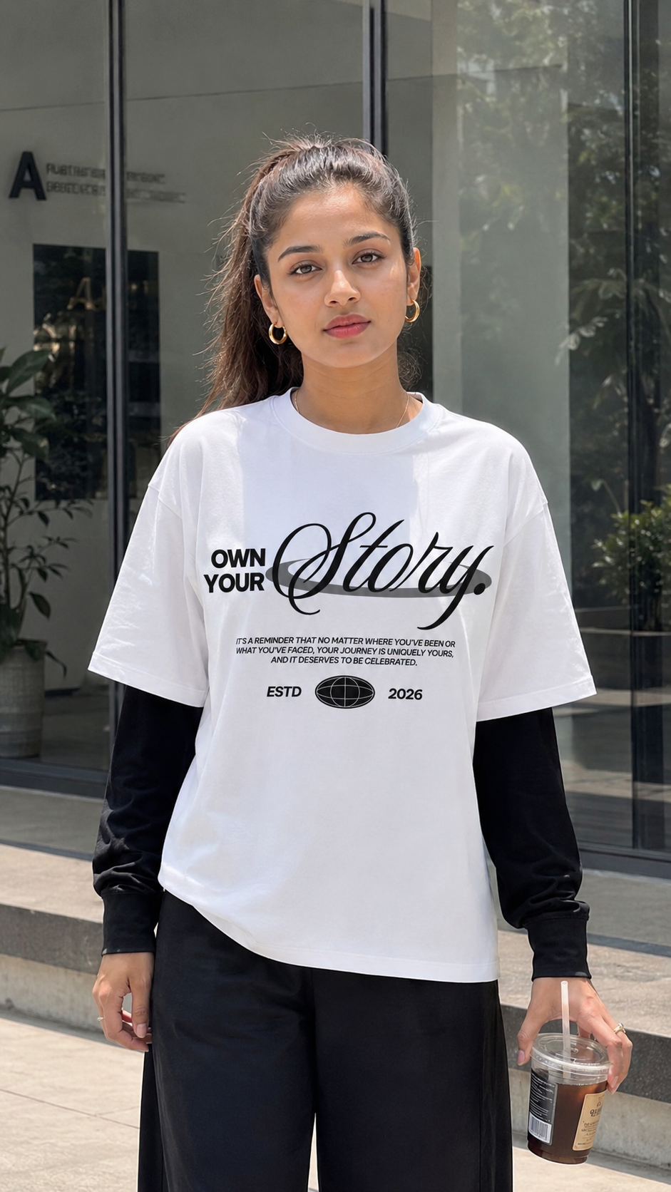 A woman wearing a Scrollfit white graphic t-shirt with black text that reads 'OWN YOUR Story' and 'ESTD 2026', layered over a black long-sleeve shirt and black pants, holding an iced coffee in front of a modern glass building.