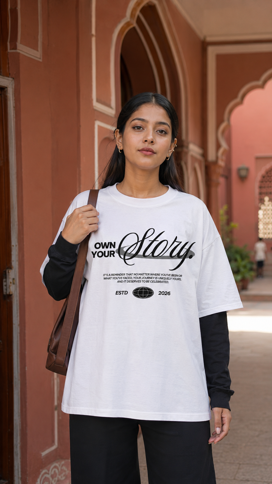 A woman wearing an oversized Scrollfit white graphic T-shirt with black text and a black long-sleeve undershirt, standing in front of a terracotta-colored archway. She is carrying a brown shoulder bag.