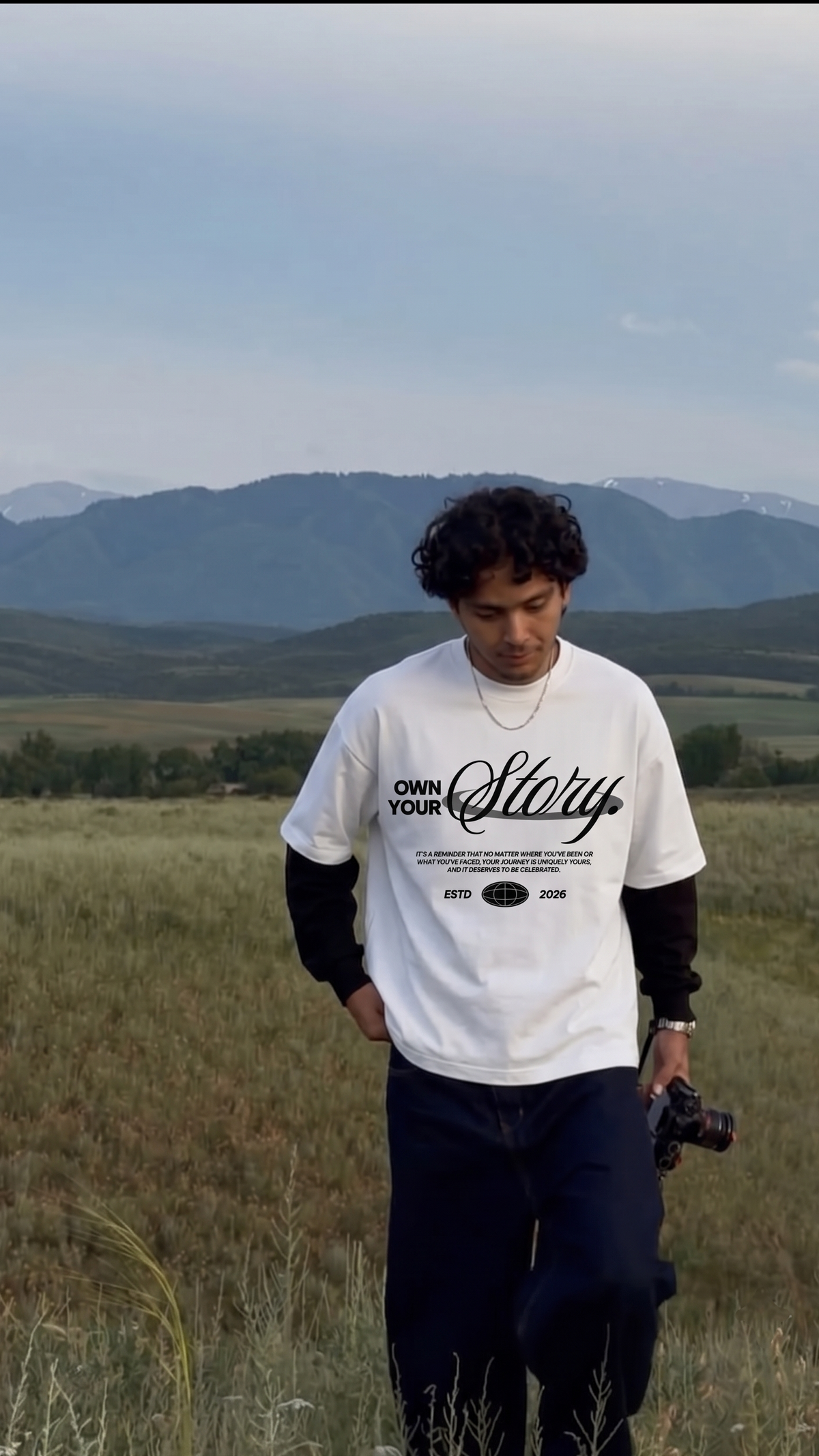 A man standing in an open grassy field with mountains in the background, wearing a Scrollfit white graphic T-shirt over a black long-sleeve shirt and dark pants.