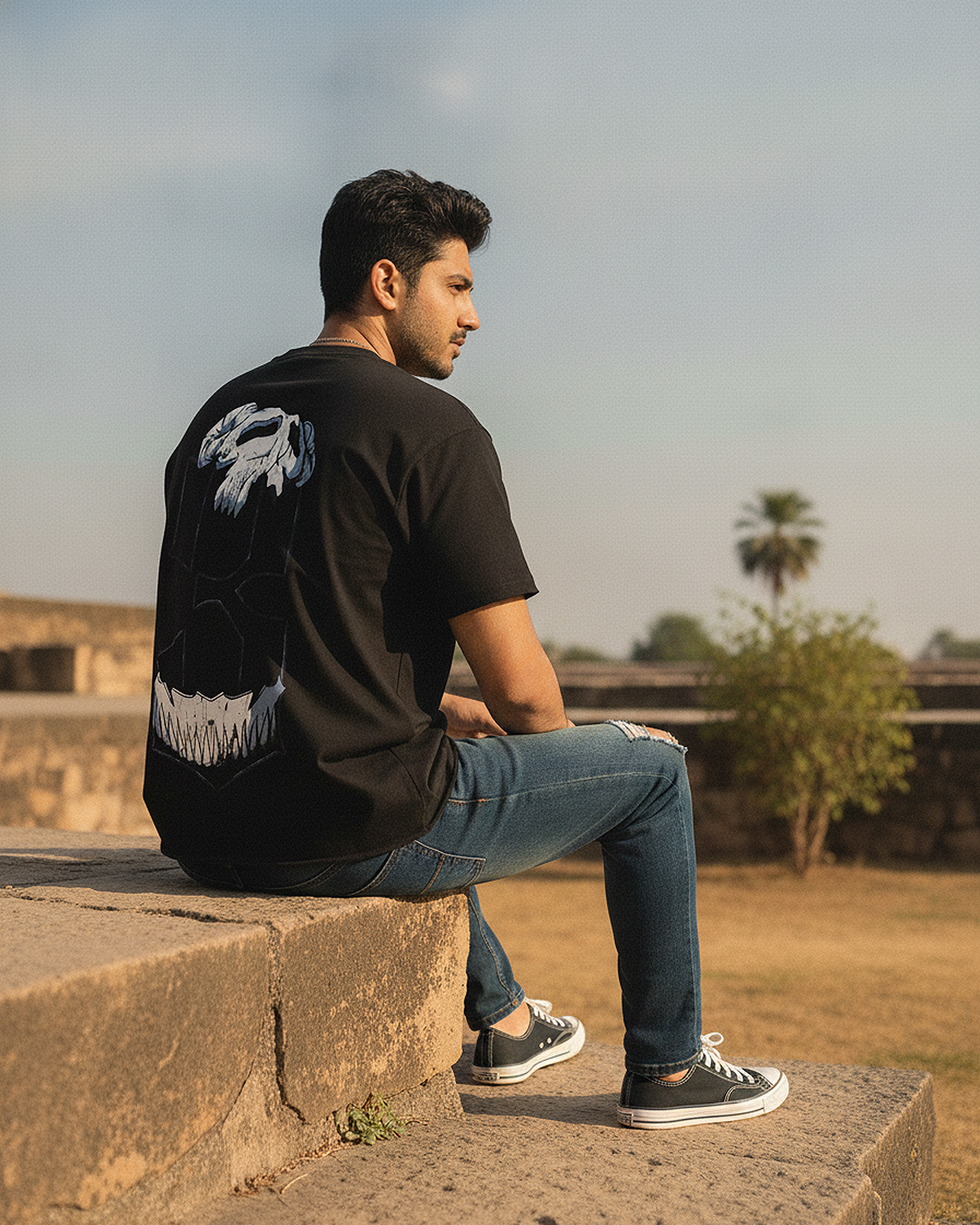 Man sitting on a stone wall wearing a The Third Graphic Boxy Fit T-Shirt with a white design on the back, in an outdoor setting.