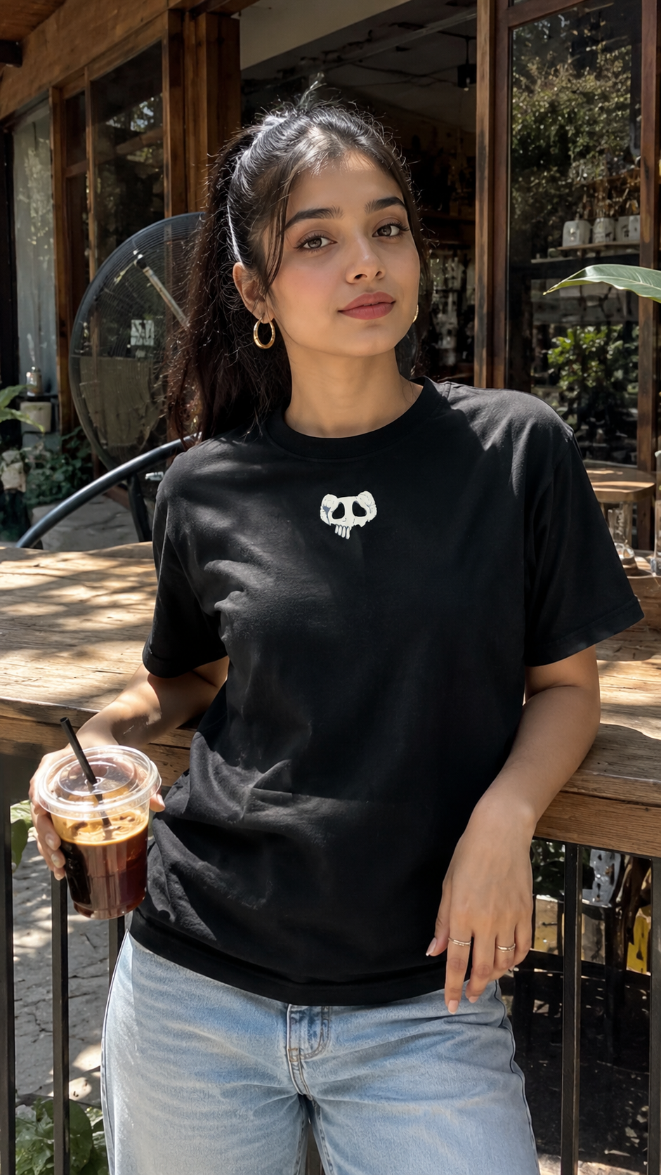 A woman wearing a Scrollfit black short-sleeve t-shirt with a small white skull graphic on the chest, paired with light blue jeans, standing at an outdoor cafe table holding an iced coffee.