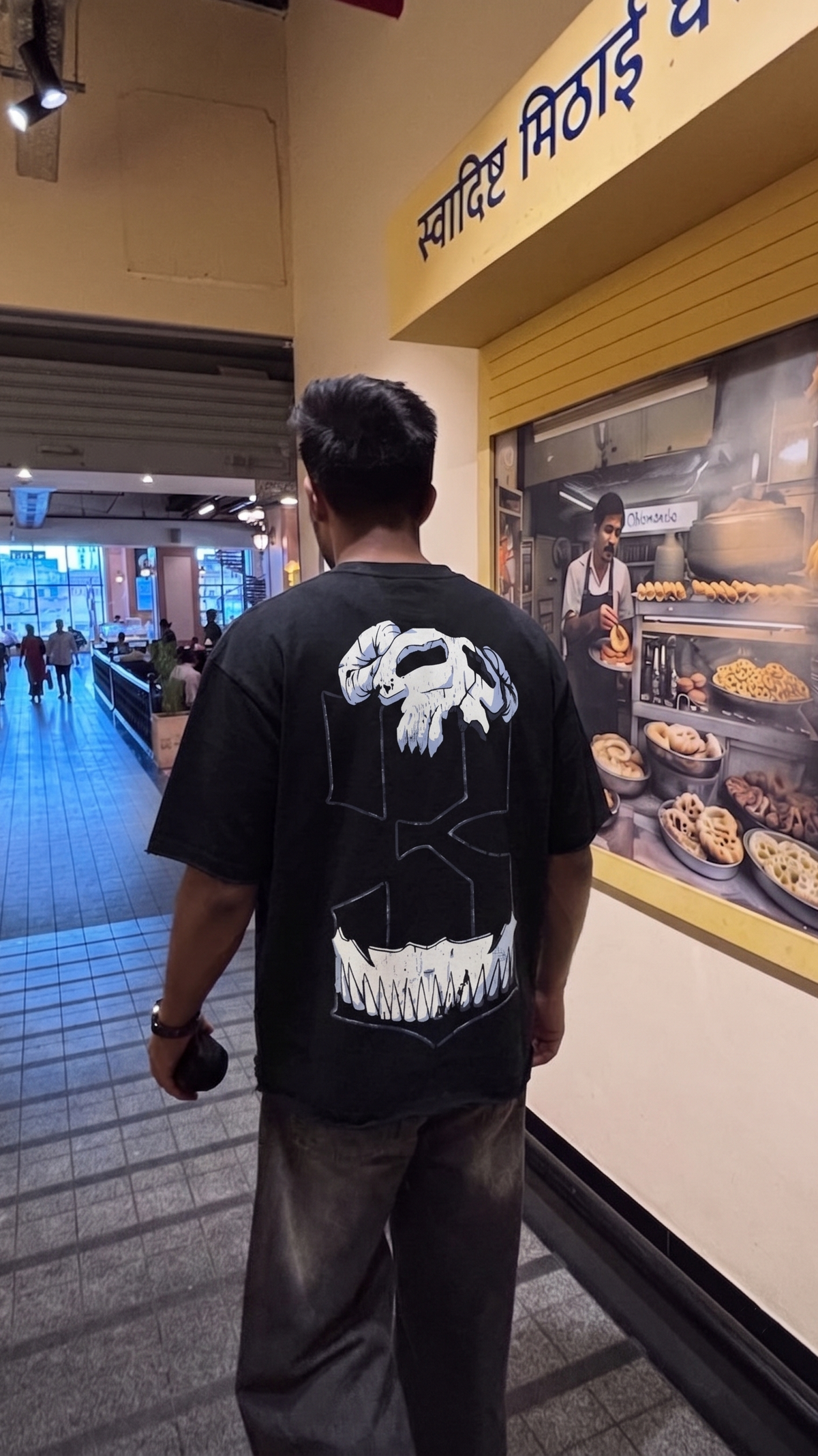 A man viewed from behind wearing a Scrollfit black graphic t-shirt with a large white skull and teeth design on the back, standing in a food court or market hallway next to a shop with Hindi signage.